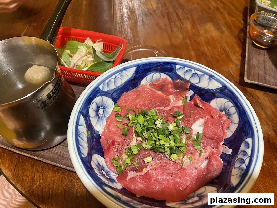 A bowl of raw beef with scallions, a pot of broth, and fresh greens on a wooden table.