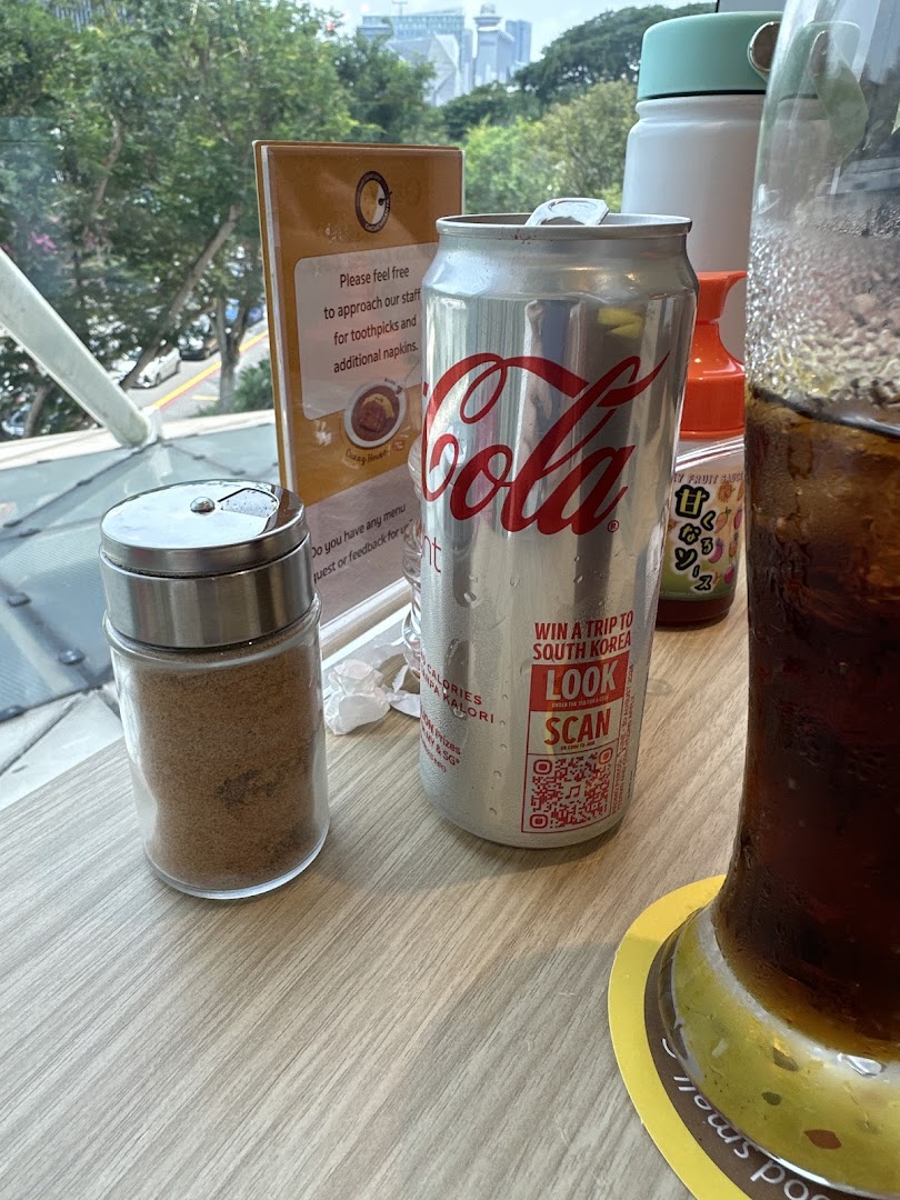 A table setting at CoCo ICHIBANYA featuring a Coca-Cola can, condiment shaker, and a window view.