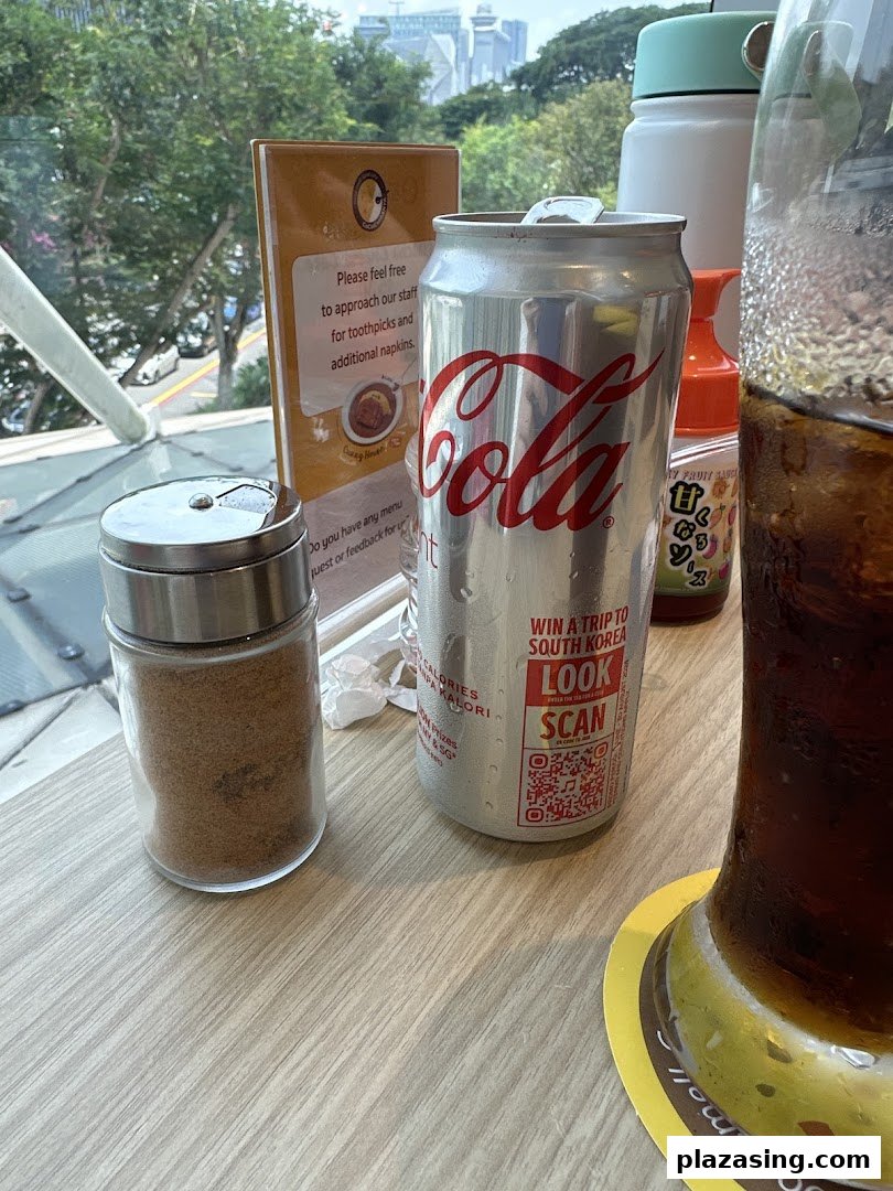 A table setting at CoCo ICHIBANYA featuring a Coca-Cola can, condiment shaker, and a window view.