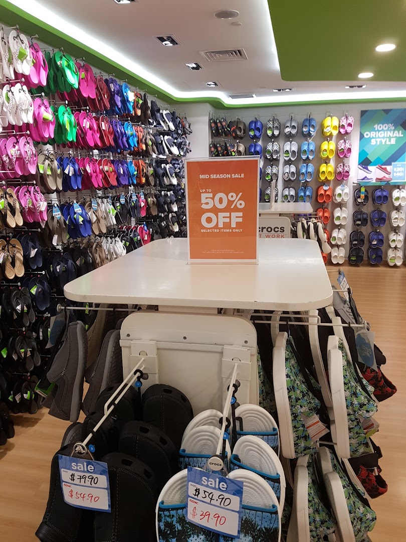 An interior view of a Crocs store showcasing colorful footwear displays and a prominent mid-season sale sign.