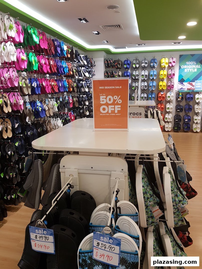 An interior view of a Crocs store showcasing colorful footwear displays and a prominent mid-season sale sign.