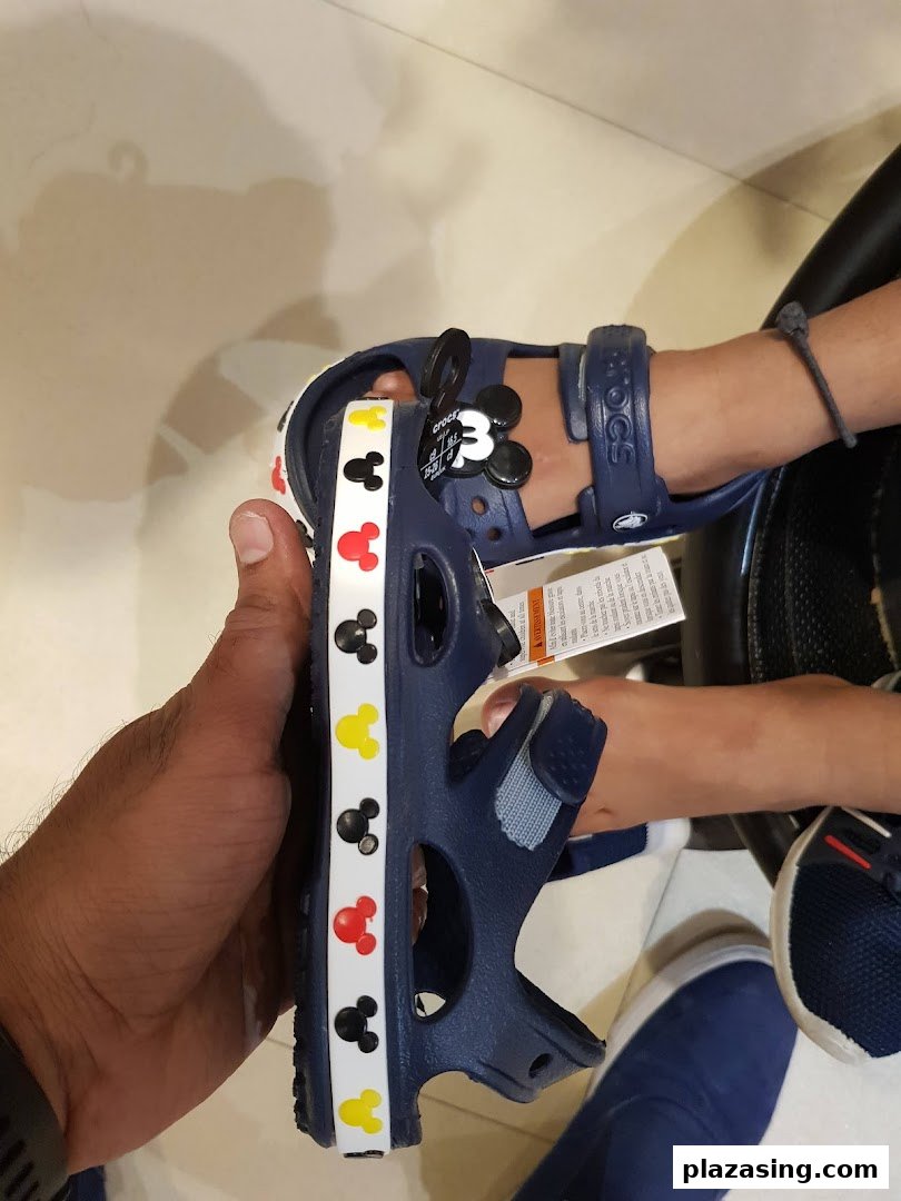 A hand holds a blue Crocs sandal with Mickey Mouse designs, next to a child's foot wearing a similar pair.