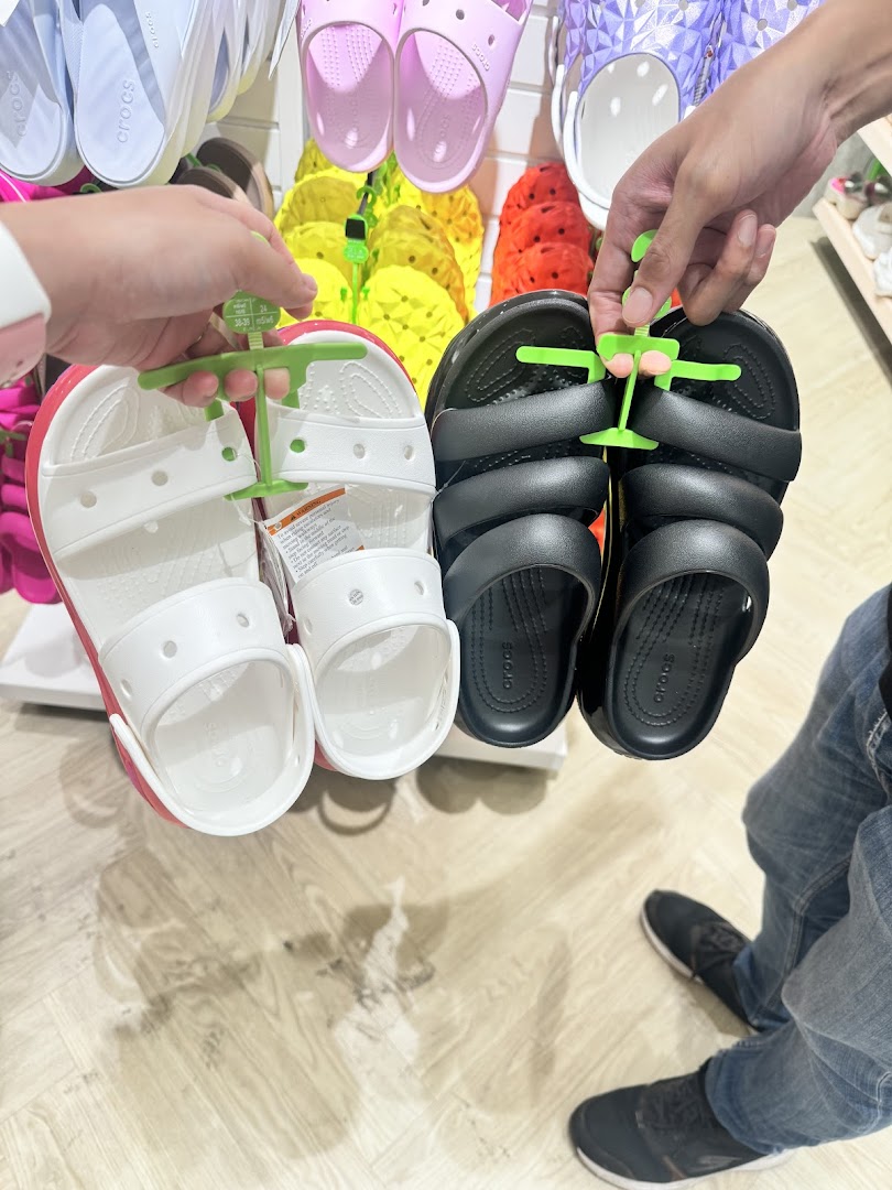 Two people hold up different styles of Crocs sandals in a vibrant store setting.