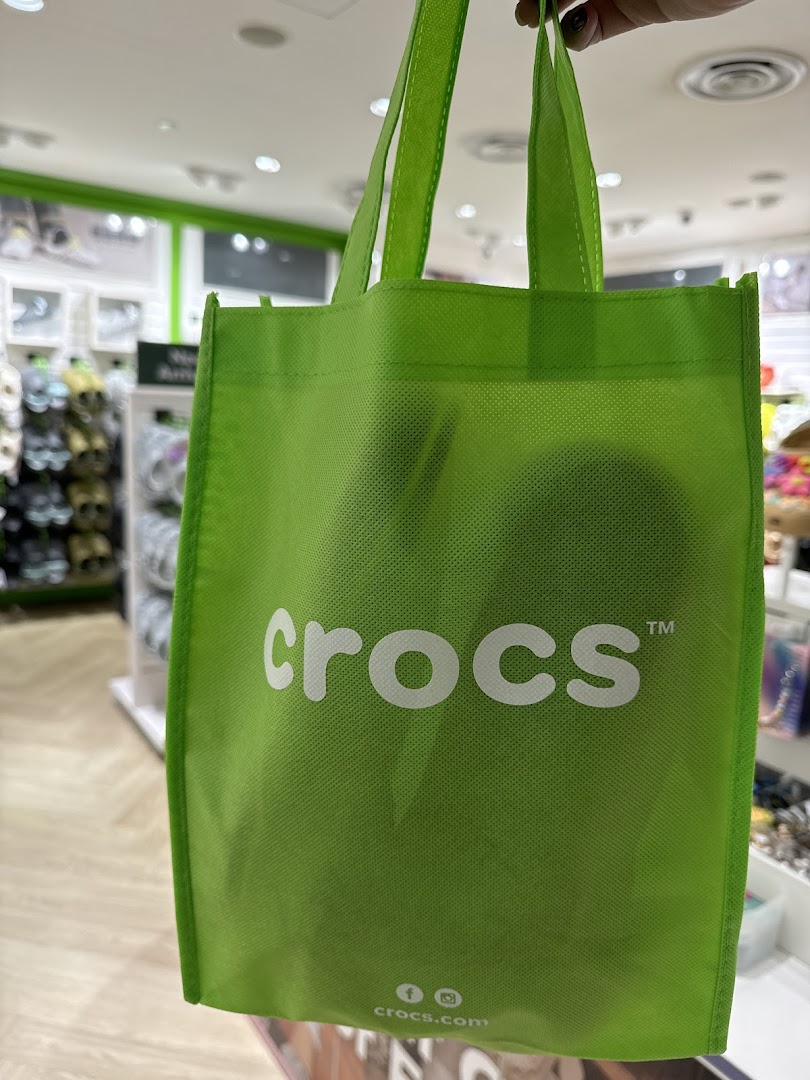 A bright green Crocs shopping bag held inside the Crocs store, showcasing the brand logo.