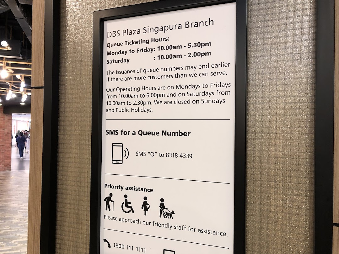 A sign displaying queue ticketing and operating hours, SMS queue number details, and priority assistance information for DBS Plaza Singapura Branch.