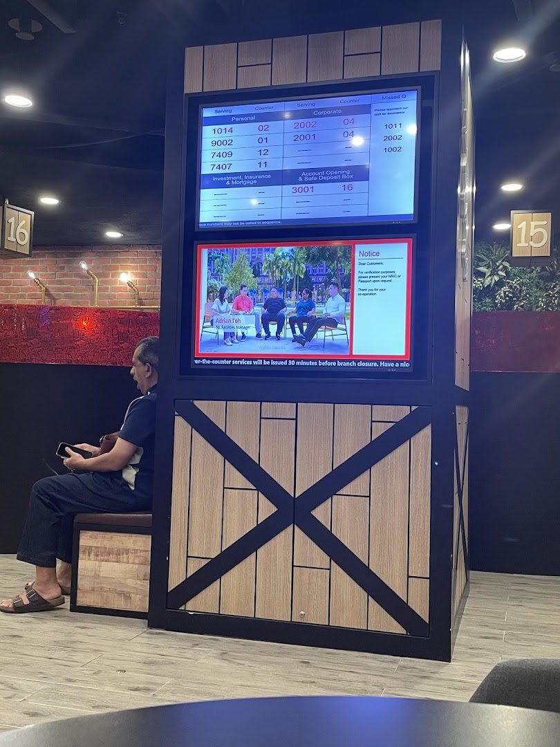 A customer waits in a modern banking hall with a digital queue management system displaying service numbers.