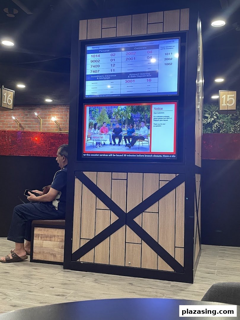 A customer waits in a modern banking hall with a digital queue management system displaying service numbers.