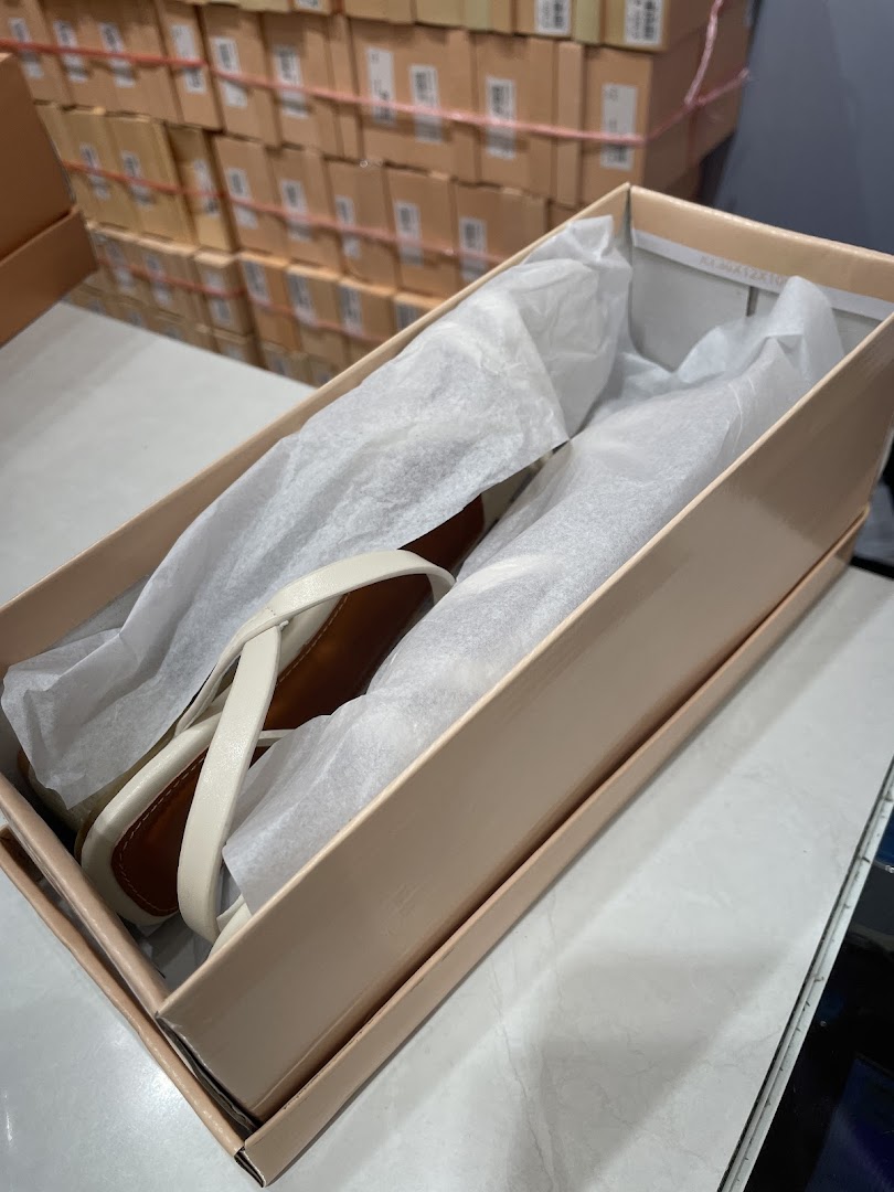 A pair of elegant white sandals in an open shoebox, surrounded by stacked inventory boxes.