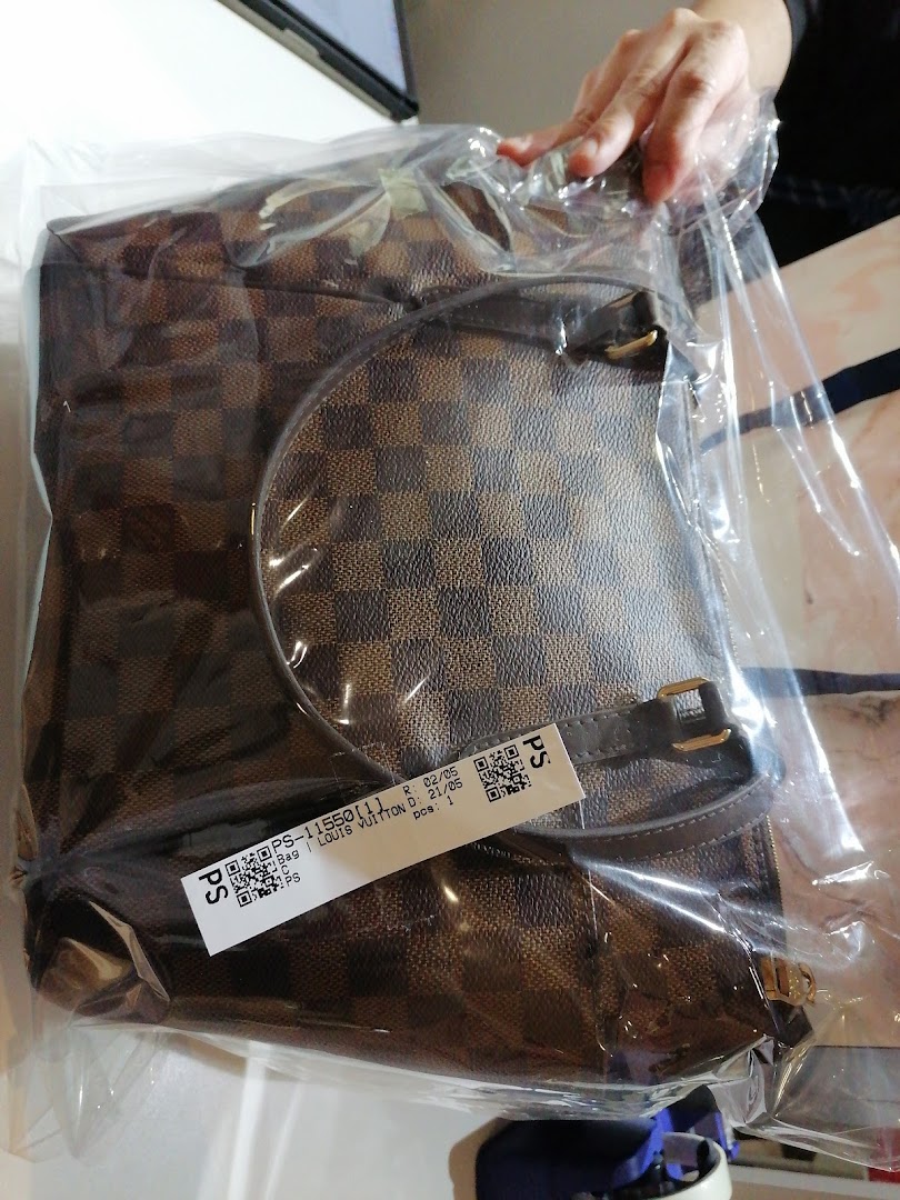 A brown checkered bag, possibly Louis Vuitton, sealed in plastic with a label, being handled.