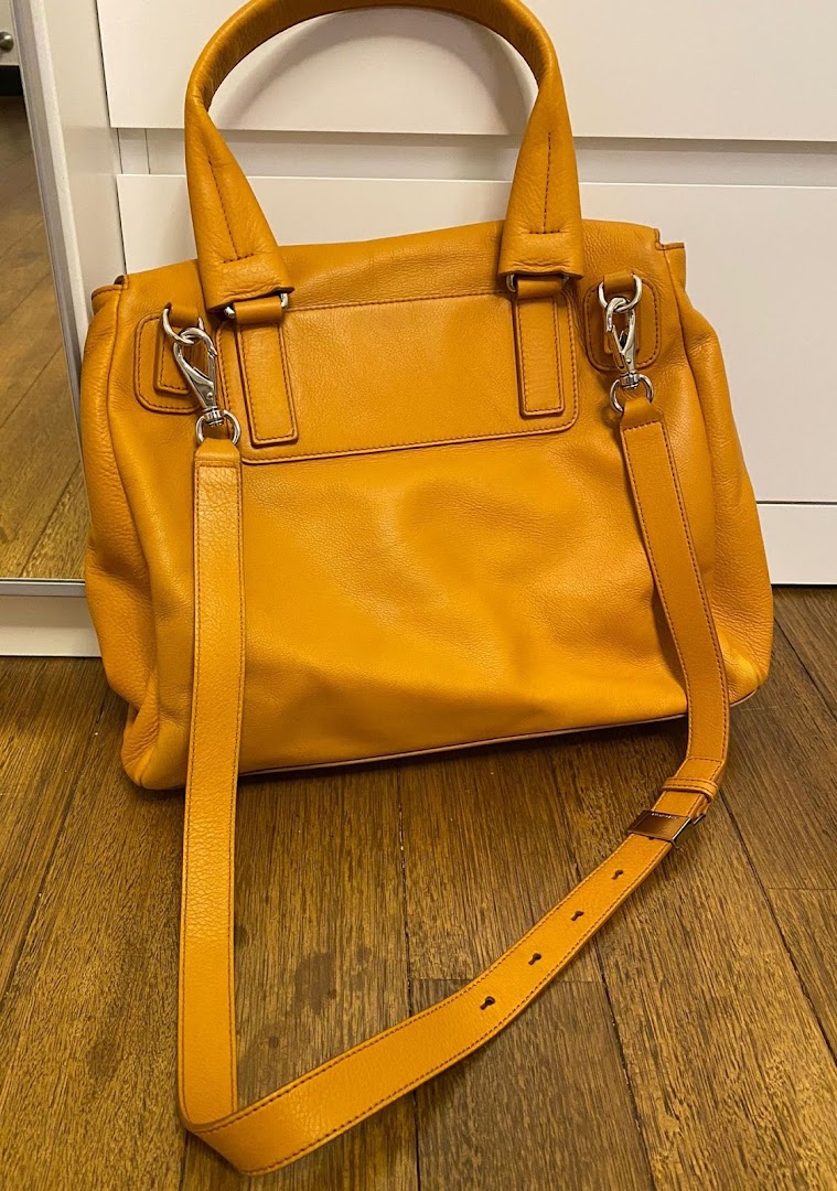 A vibrant orange leather handbag with a detachable strap displayed on a wooden floor.