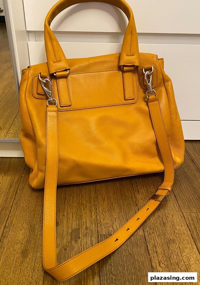 A vibrant orange leather handbag with a detachable strap displayed on a wooden floor.