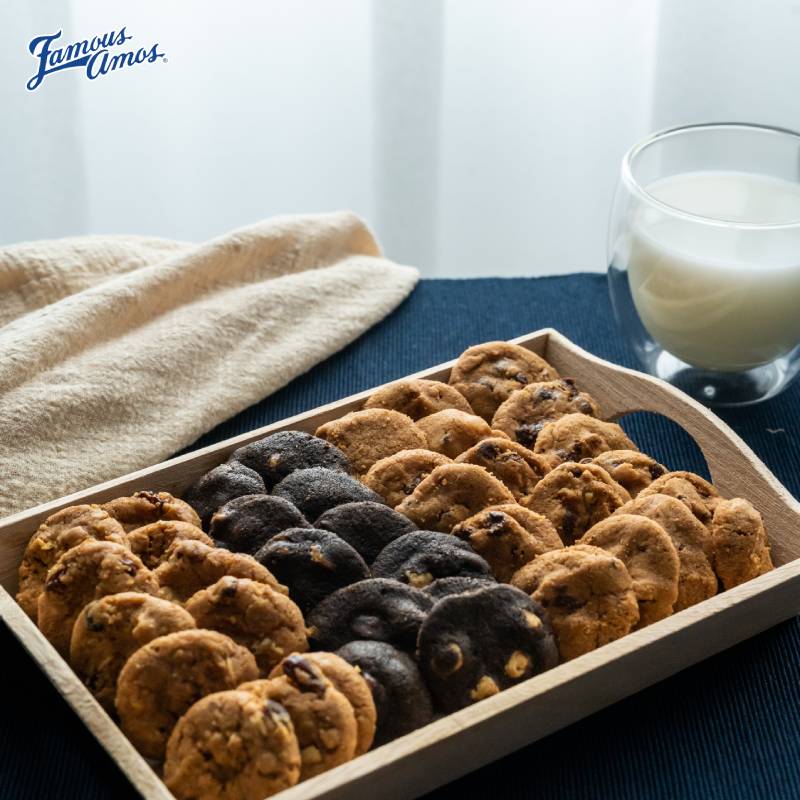 A wooden tray filled with an assortment of freshly baked Famous Amos cookies, accompanied by a glass of milk.