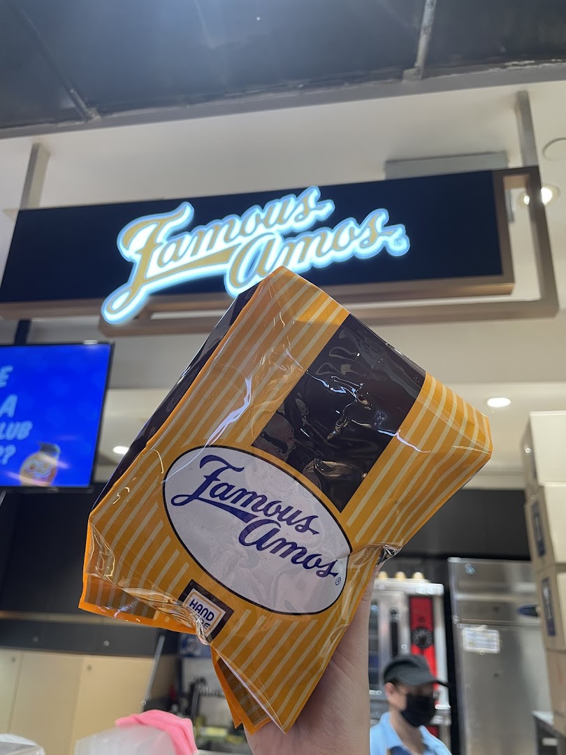 A hand holds a Famous Amos cookie bag in front of the illuminated shop sign.