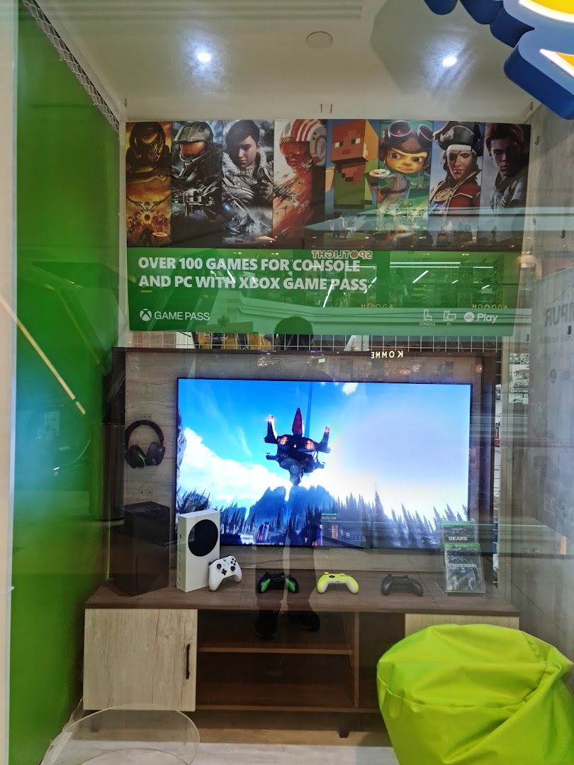 A vibrant gaming display featuring Xbox consoles, controllers, a large TV, and game posters.