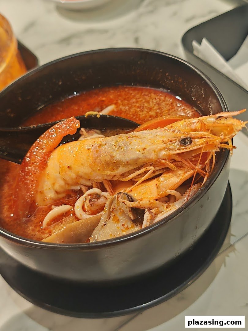 A close-up of a vibrant seafood tom yum soup with large prawns, squid, and mussels in a black bowl.