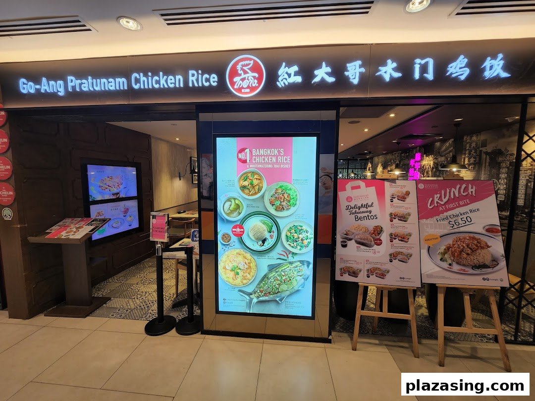 The brightly lit storefront of Go-Ang Pratunam Chicken Rice, showcasing its entrance and promotional displays.