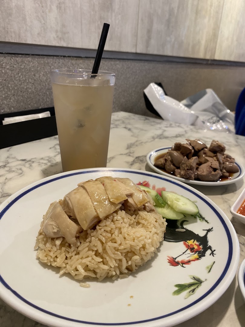 A plate of Hainanese chicken rice with sliced chicken and cucumber, served with a drink and a side dish.