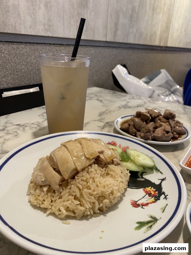 A plate of Hainanese chicken rice with sliced chicken and cucumber, served with a drink and a side dish.