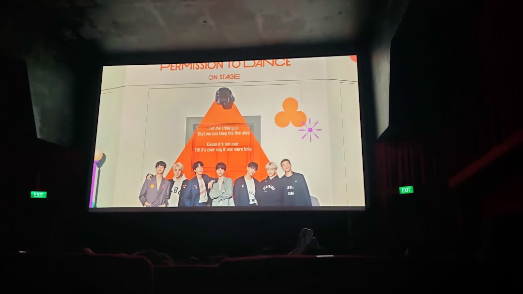 A cinema screen displaying 'Permission to Dance On Stage' content to an audience in a dark theater.