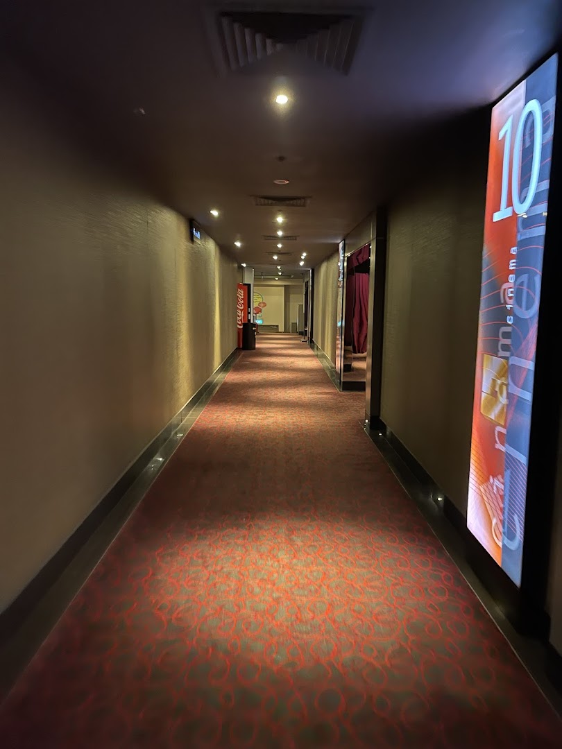 A long, dimly lit hallway with red patterned carpet, leading to cinema halls, featuring a Coca-Cola machine and a hall number sign.