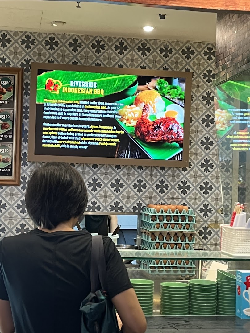 A person stands at a food hall counter with a digital menu board displaying Indonesian BBQ dishes.