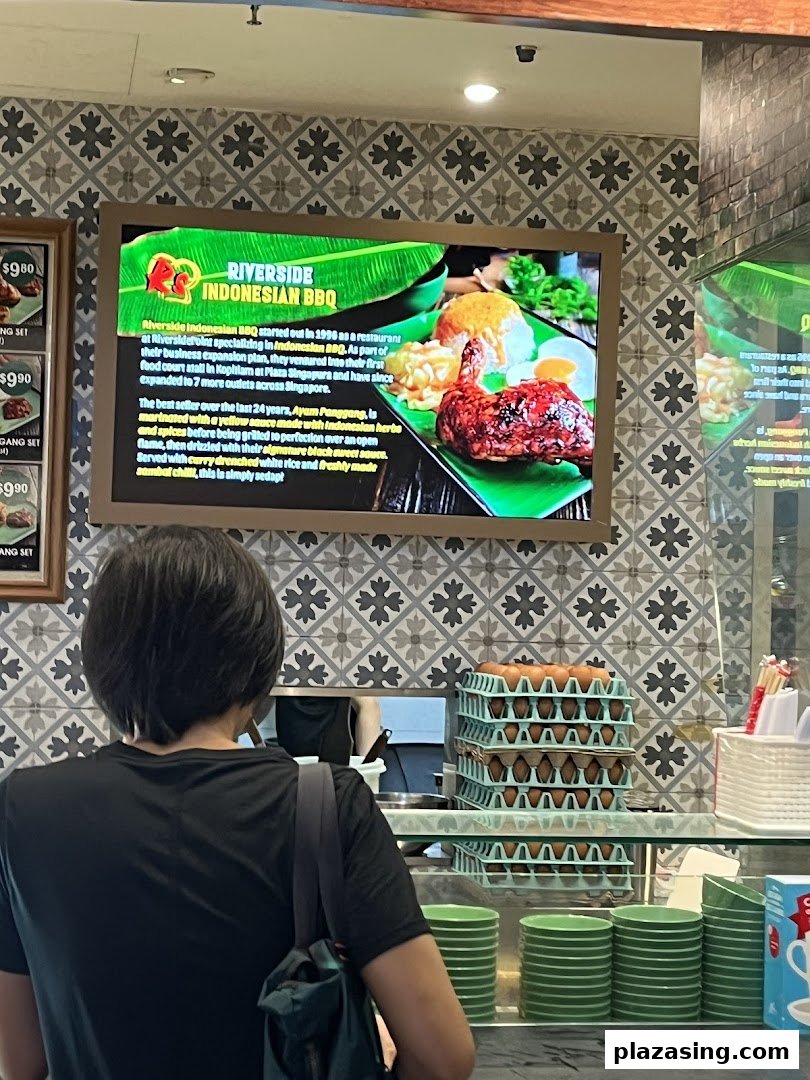 A person stands at a food hall counter with a digital menu board displaying Indonesian BBQ dishes.