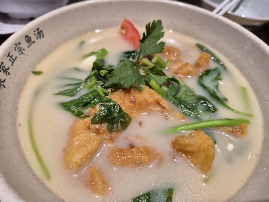 A close-up of a creamy fish soup with fried fish pieces, green vegetables, and a tomato slice.