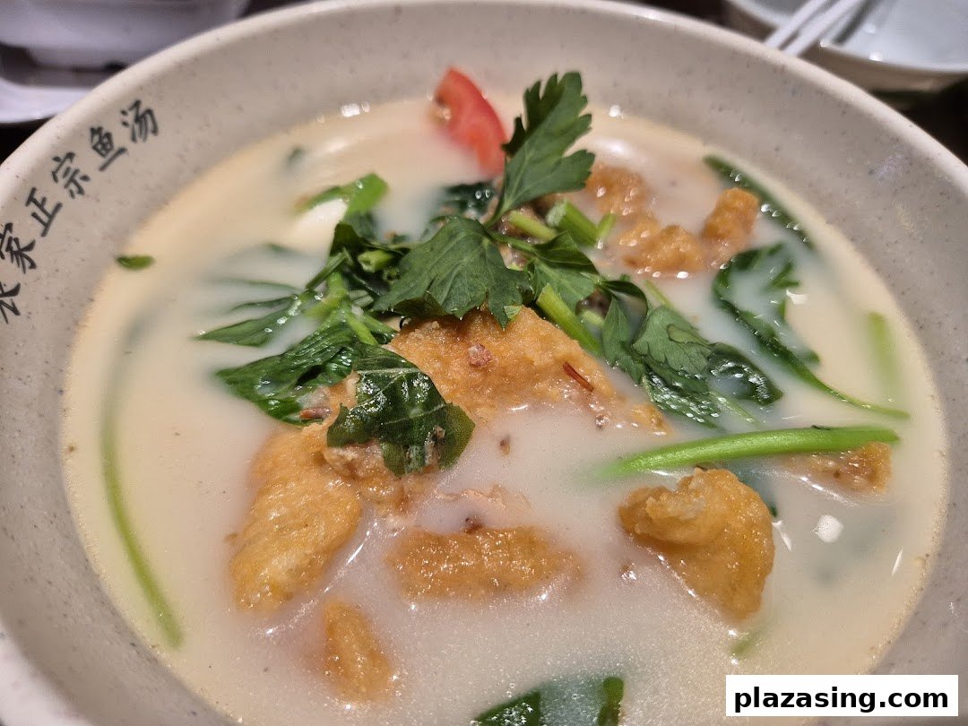 A close-up of a creamy fish soup with fried fish pieces, green vegetables, and a tomato slice.