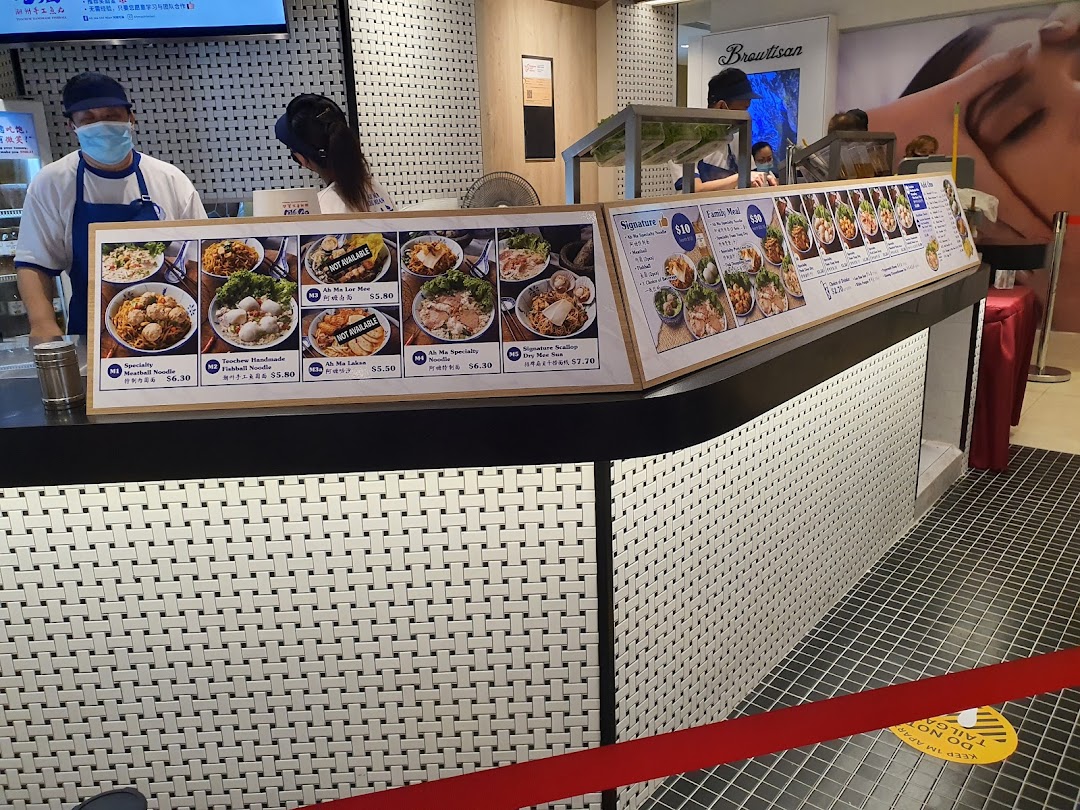 A food stall counter at Kopitiam Food Hall displaying various noodle dishes and prices on large menu boards.