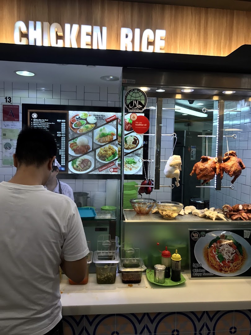 A customer at a Chicken Rice stall in Kopitiam Food Hall, featuring hanging roasted chickens and a digital menu.