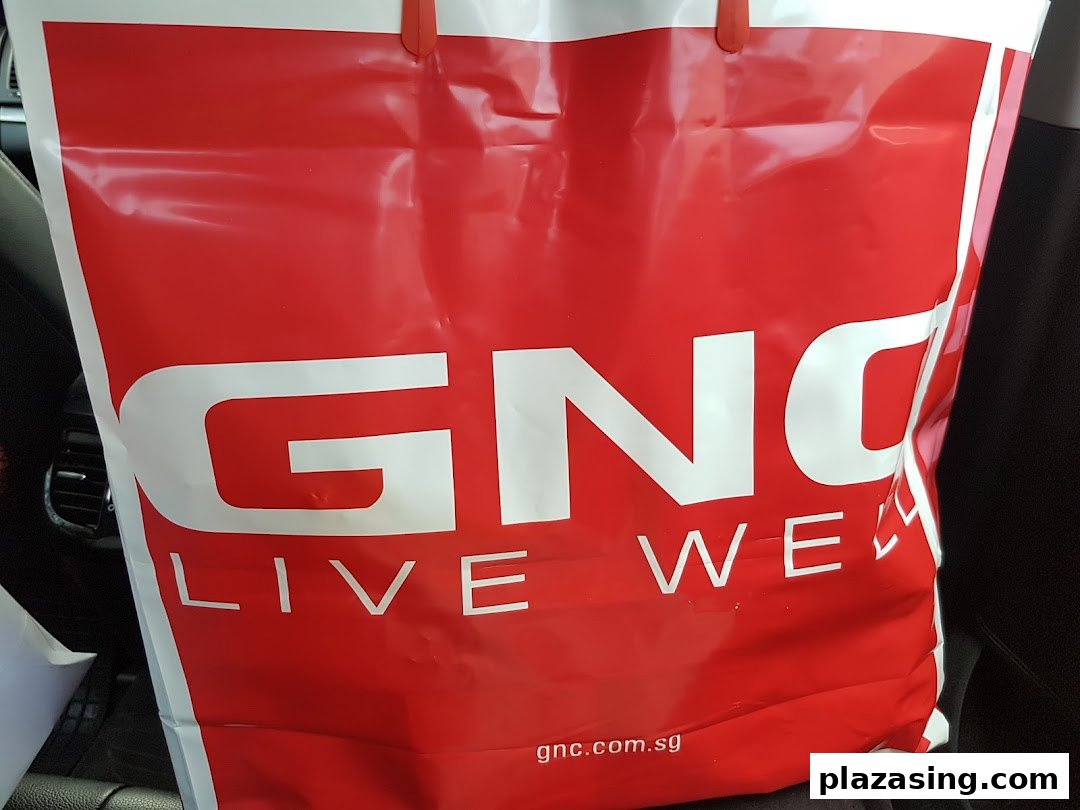 A vibrant red GNC shopping bag featuring the 'LIVE WELL' slogan and brand website.