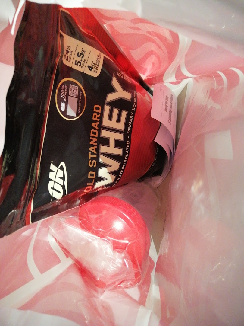 A shopping bag containing Optimum Nutrition Gold Standard Whey protein and a red shaker bottle.