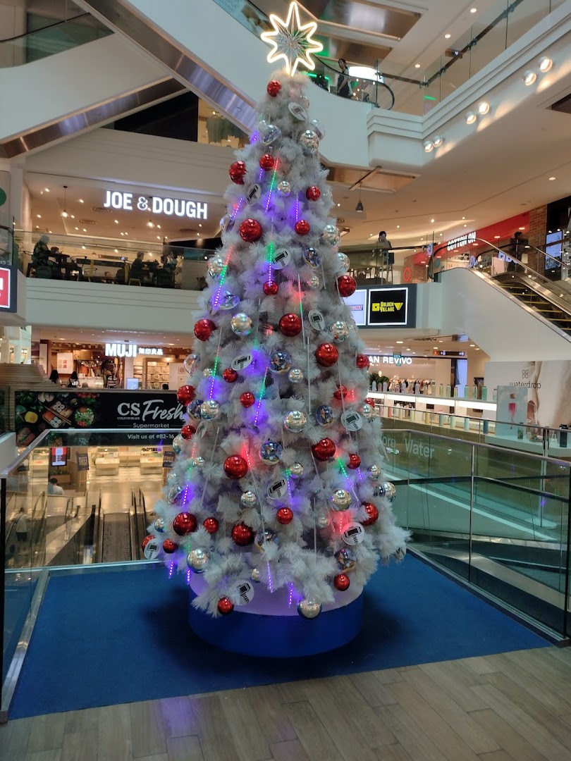 A festive white Christmas tree adorned with red and silver ornaments stands prominently in a multi-level shopping mall.
