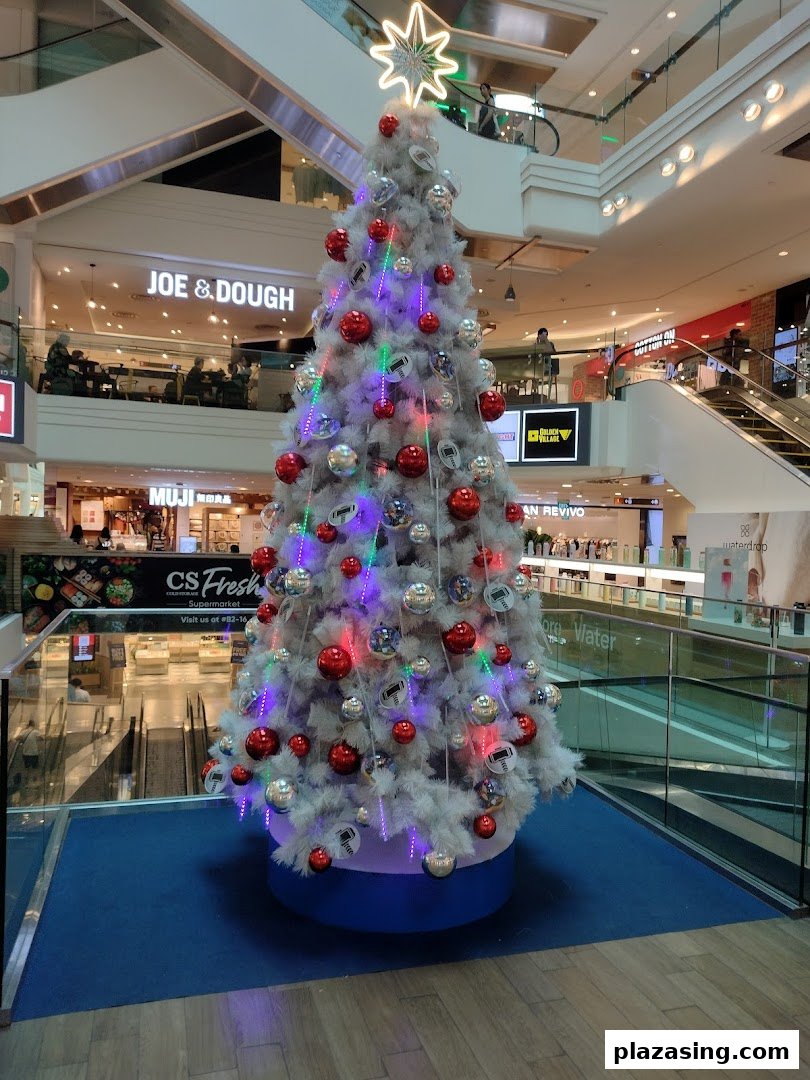 A festive white Christmas tree adorned with red and silver ornaments stands prominently in a multi-level shopping mall.