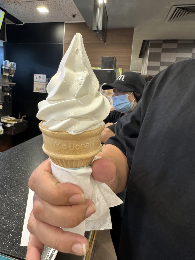 A hand holding a McDonald's soft-serve ice cream cone inside the restaurant, with a staff member in the background.