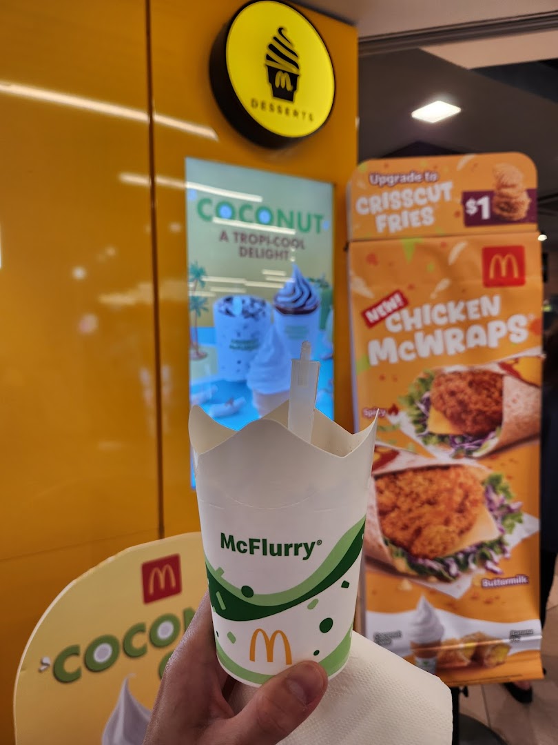 A hand holds a McFlurry cup in front of McDonald's promotional displays for desserts and new wraps.