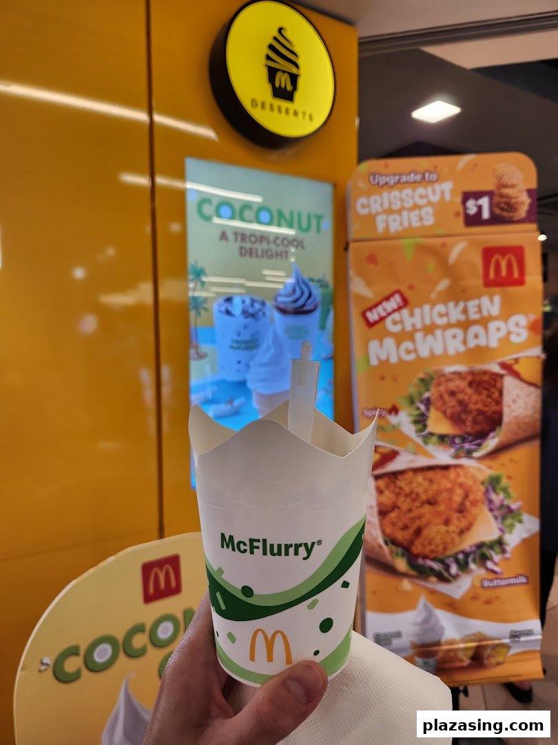 A hand holds a McFlurry cup in front of McDonald's promotional displays for desserts and new wraps.
