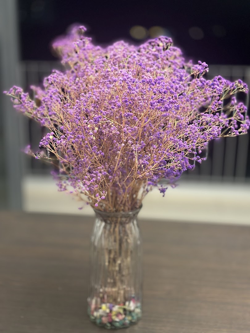 A vase filled with delicate purple dried flowers sits on a dark wooden table.