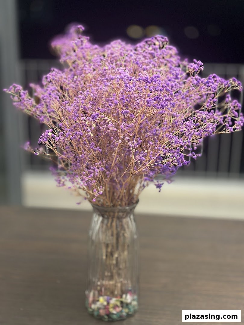 A vase filled with delicate purple dried flowers sits on a dark wooden table.