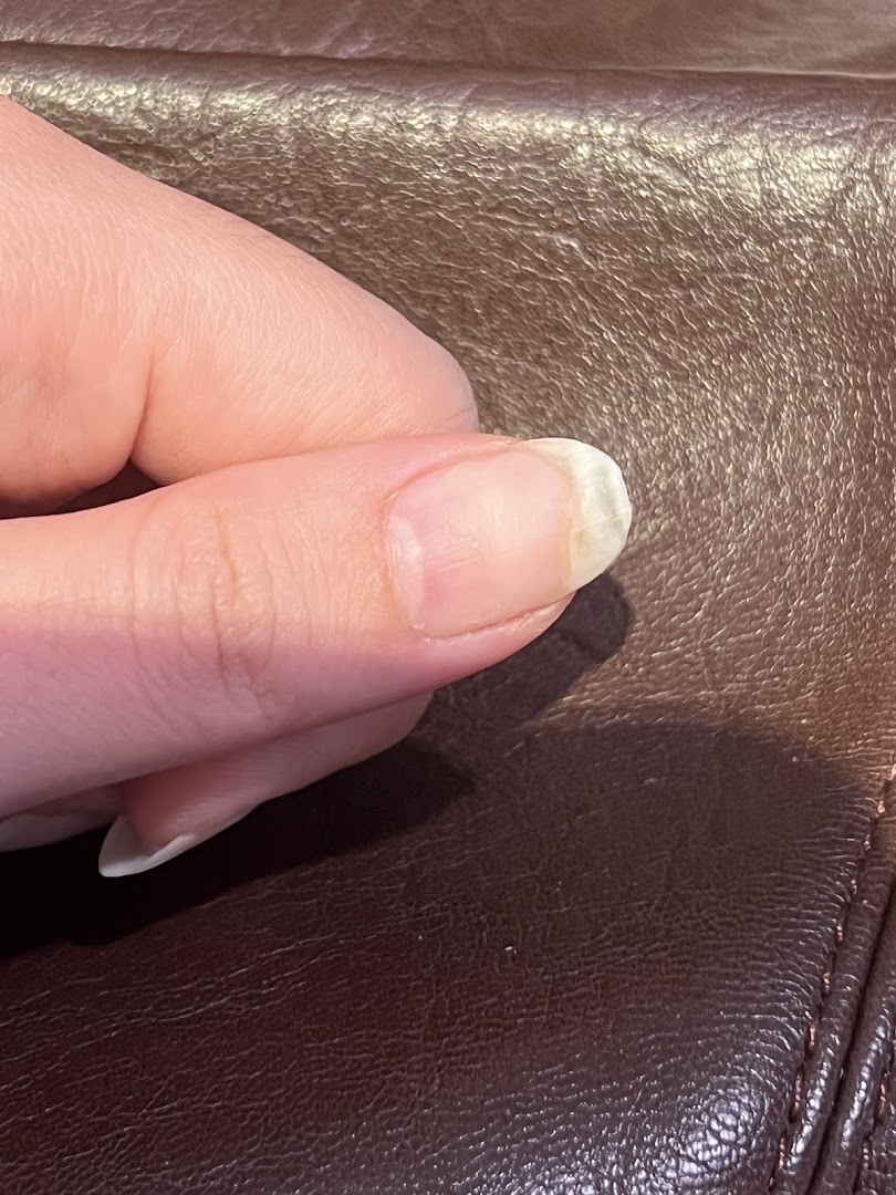 A close-up of a thumb and fingernail, resting on a textured brown surface, likely at the nail spa.