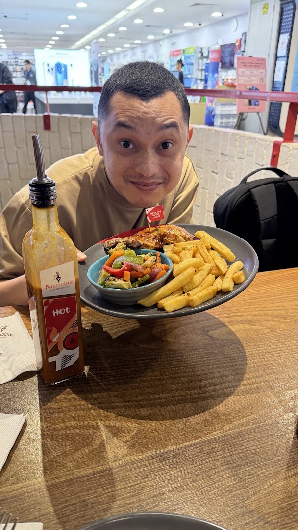 A person smiling while holding a plate of Nando's flame-grilled chicken with sides and hot sauce.