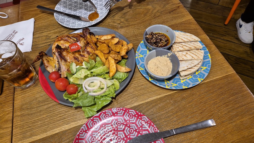 A flame-grilled PERi-PERi chicken meal with wedges, salad, hummus, and pita bread on a wooden table.