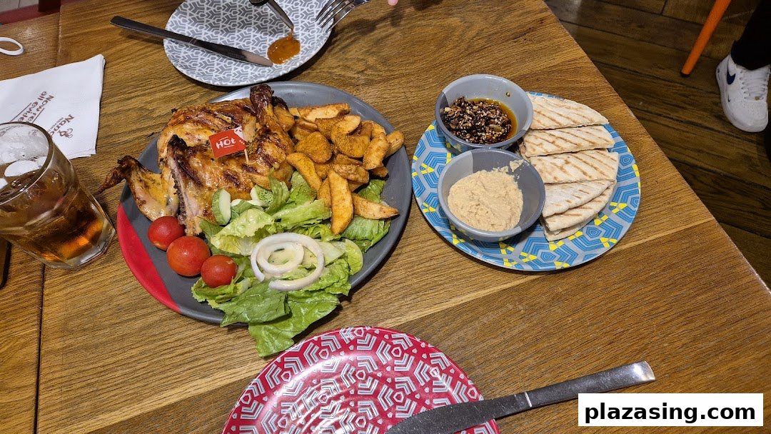 A flame-grilled PERi-PERi chicken meal with wedges, salad, hummus, and pita bread on a wooden table.