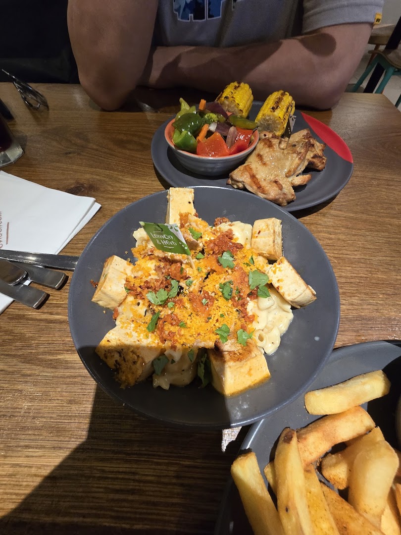 Plates of Nando's flame-grilled chicken, mac and cheese with tofu, vegetables, corn, and fries on a wooden table.