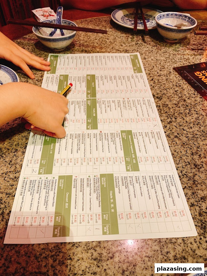 A person's hands are seen pointing at a detailed menu on a table in a restaurant setting.