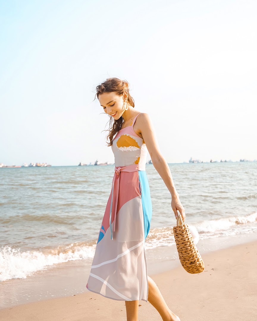 A woman in a vibrant, patterned dress walks along a sunny beach with the ocean in the background.