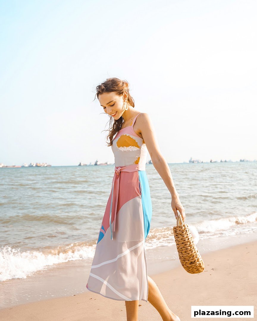 A woman in a vibrant, patterned dress walks along a sunny beach with the ocean in the background.