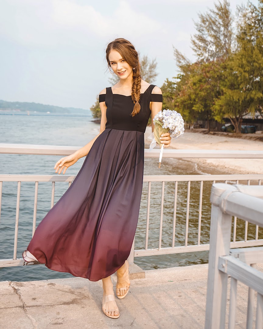 A woman in an elegant off-shoulder ombre dress holding flowers on a pier by the water.
