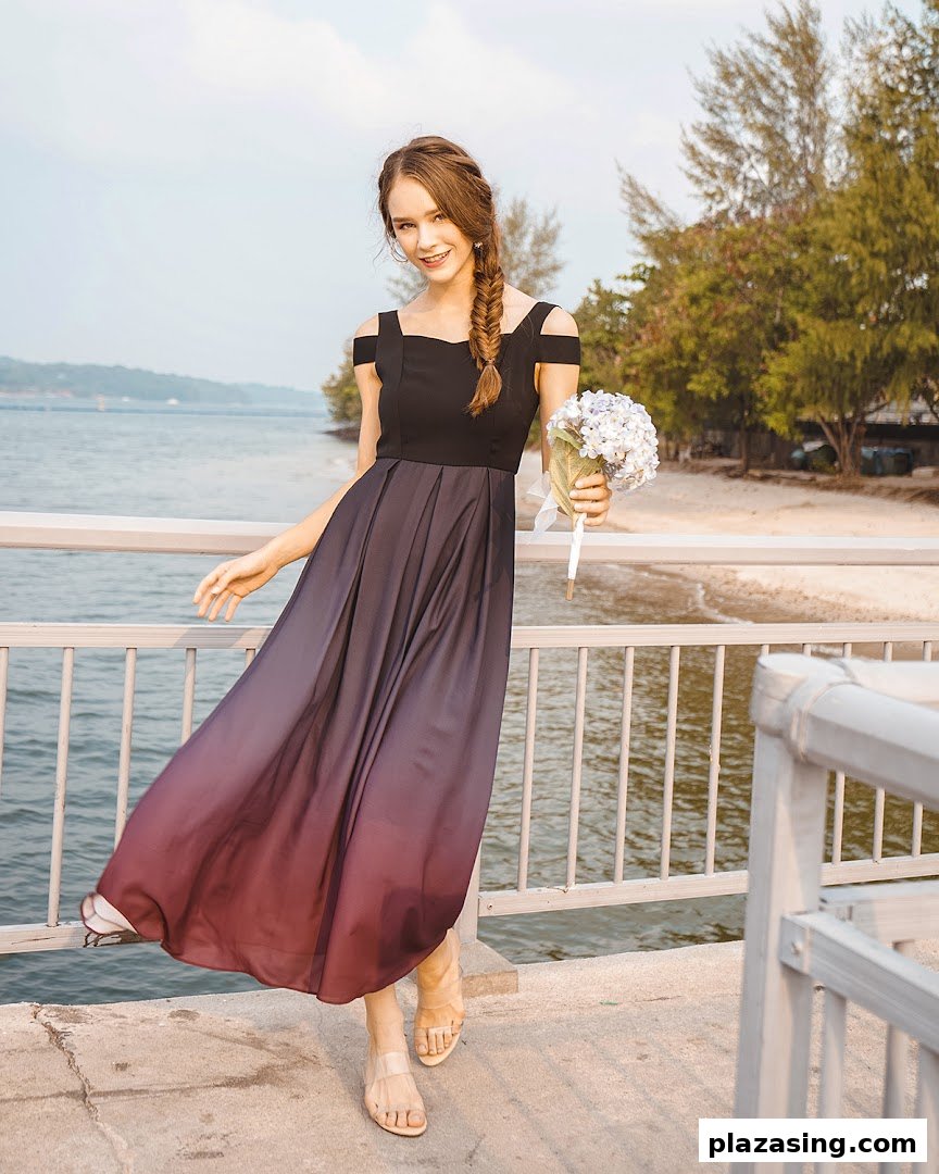 A woman in an elegant off-shoulder ombre dress holding flowers on a pier by the water.