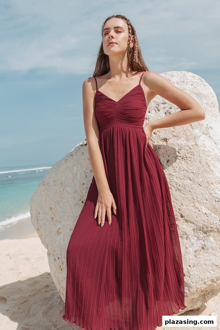 A model in a flowing maroon pleated dress poses by a rock on a sunny beach.